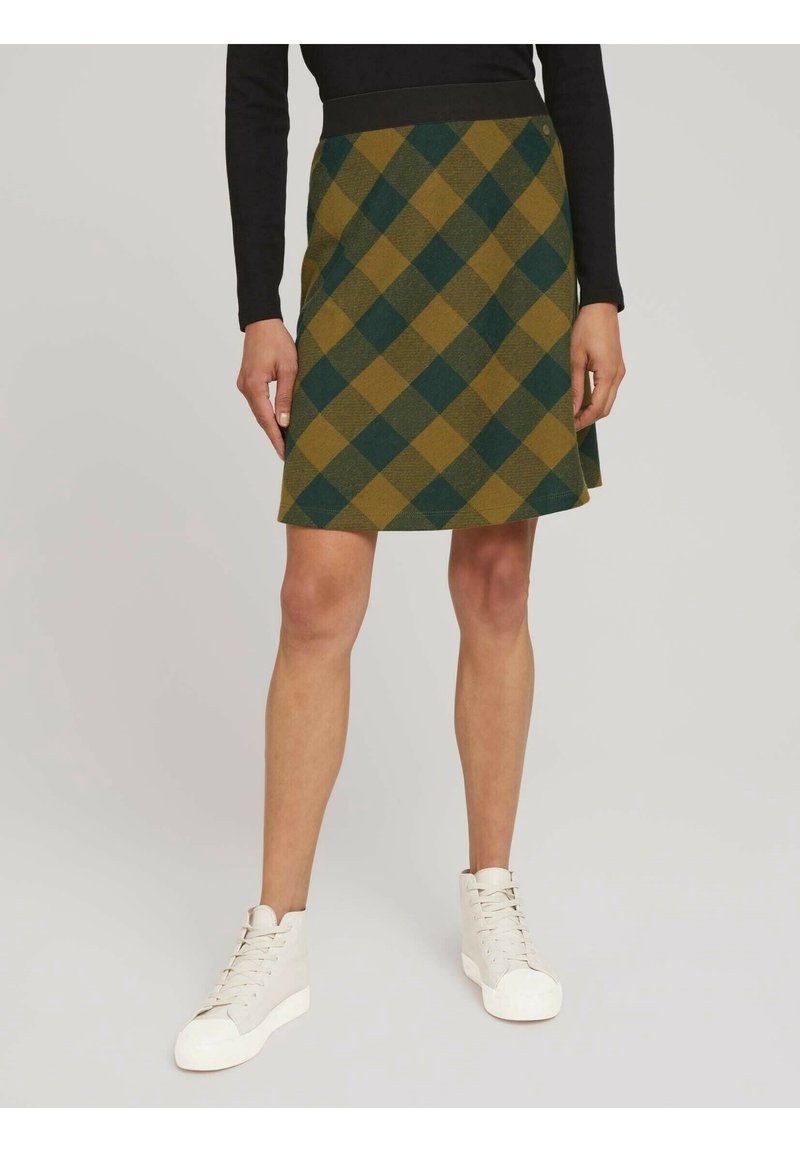 Femme portant une jupe à motifs losanges verts et moutarde, avec un haut noir et des baskets montantes blanches, debout sur un sol gris clair.