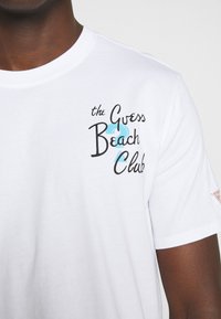 Weißes Baumwoll-T-Shirt mit "the Guess Beach Club" in Schwarz und Türkis gestickt. Crewneck und kurze Ärmel. Einfaches Design.