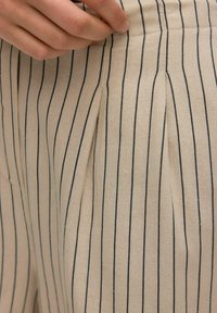 Beige plisserede bukser lavet af bomuldsstof, med lodrette marineblå striber, en synlig talje og en hånd, der griber fat om stoffet.