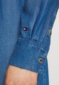 Manica di camicia in denim blu con chiusura a bottone, caratterizzata da un piccolo logo ricamato in rosso, bianco e blu sul polsino. Tessuto texturizzato.
