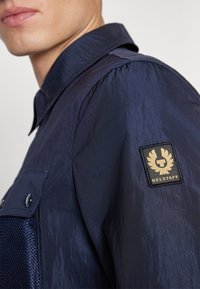 Detailní záběr na osobu v tmavě modré bundě s logem Belstaff na horním rukávu a knoflíkovou náprsní kapsou.
