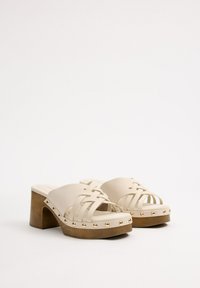 Mules de tacón alto beige con diseño superior trenzado, que presentan tachuelas doradas a lo largo de la suela y un tacón de bloque de madera. Textura suave.