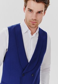 Granatowa, dwurzędowa kamizelka z wzorzystej tkaniny, z kołnierzem szalowym i dwoma guzikami z przodu, noszona na białej koszuli.