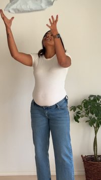 Balta prigludusi marškinėliai, mėlyni džinsai ir pakeltos rankos, laikantys sidabrinį paketą. Šviesi vidaus aplinka su žaliu augalu pintame vazonėlyje.