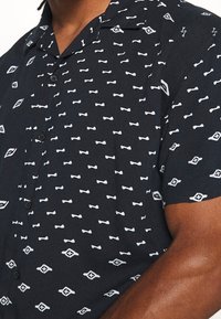 Kortärmad, kragad skjorta med marinblå bakgrund. Har vita mönster som liknar rosetter och diamanter. Knappar är synliga fram.