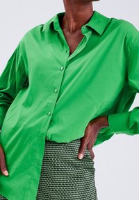 Zielona koszula z guzikami i kołnierzykiem; wykonana z gładkiego materiału, posiada długie rękawy i luźny krój. Noszona z wzorzystą spódnicą.