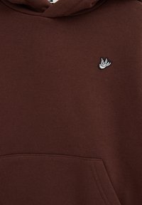 Sweat à capuche marron avec un petit emblème d'oiseau blanc brodé sur le haut de la poitrine et une poche kangourou avant visible en bas.