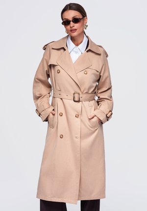 Femme portant un trench-coat beige ceinturé avec de gros boutons, une chemise blanche à col, des lunettes de soleil et des boucles d'oreilles en or, debout les mains dans les poches.