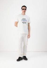 White cotton t-shirt with colorful "Tommy Jeans New York" graphic, paired with white trousers and black sneakers. Model wearing sunglasses.