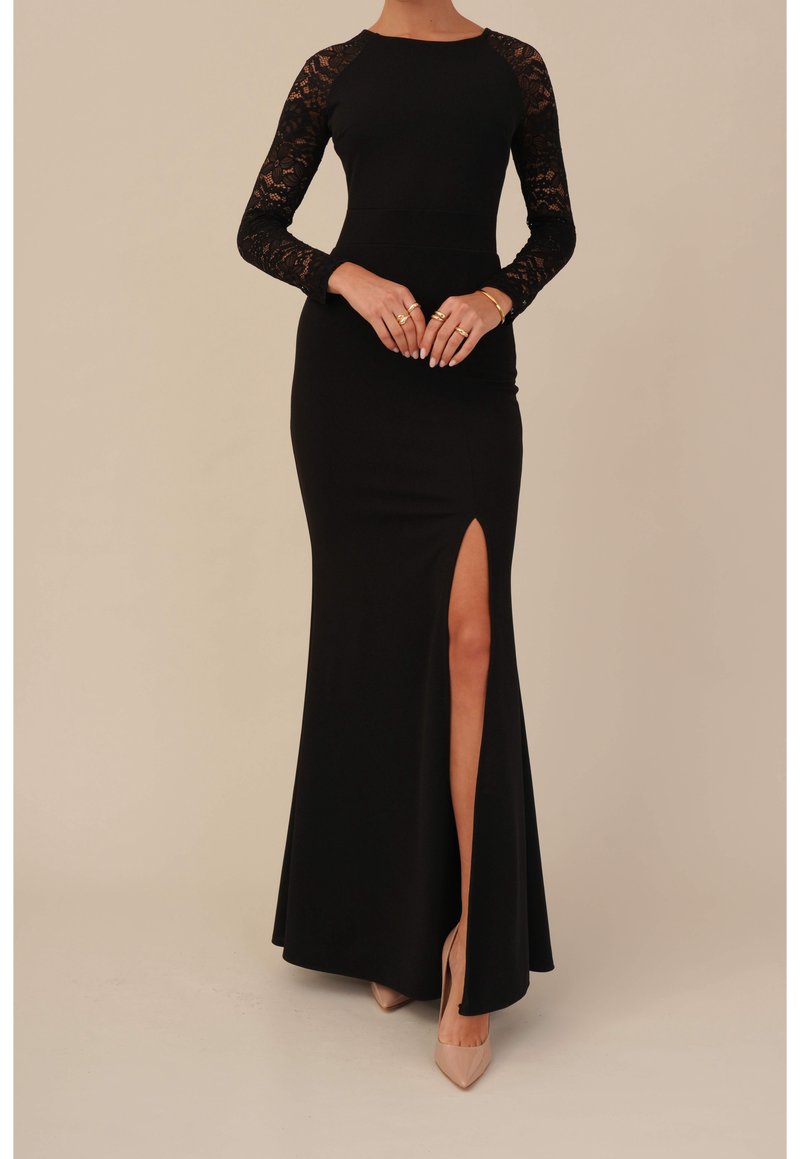 Femme portant une longue robe noire avec des manches longues en dentelle et une fente haute sur la cuisse, accessoirisée avec des bagues dorées et des talons beiges.