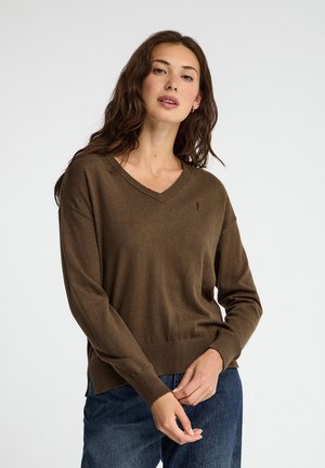 Junge Frau mit langen braunen Haaren trägt einen braunen Pullover mit V-Ausschnitt und blaue Jeans, steht vor einem schlichten hellen Hintergrund.