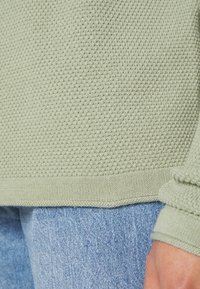 Pull en maille texturée vert clair avec un ourlet côtelé, associé à un jean en denim bleu. Gros plan mettant l'accent sur le détail du tissu.
