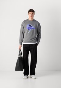 En ung mann står, iført en grå grafisk hettegenser, svarte bukser, hvite joggesko, og holder en stor svart totebag med den ene hånden.