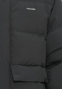 Veste doudoune noire en matériau lisse et matelassé avec un logo visible "Holzweiler". Présente un rembourrage volumineux et un détail de poche avant.