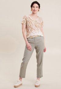 Cremfarbene V-Ausschnitt-Bluse mit goldbraunem Tierdruck, kombiniert mit hellgrünen, taillierten Hosen und weißen Sneakers mit dicker beiger Sohle.