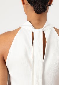 Haut halter blanc en tissu texturé. Présente une fermeture à nouer dans le dos et un design dos nu, mettant en valeur des bords lisses et des détails subtils.