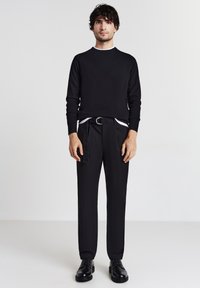 Schwarzer Strickpullover mit rundem Ausschnitt, kombiniert mit schwarzen Hosen, die einen Gürtel und seitliche Taschen haben. Schwarze Schnürschuhe vervollständigen das Outfit.