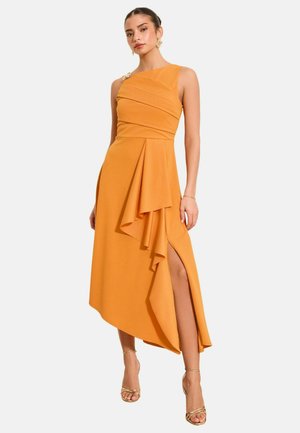 Woman wearing a sleeveless mustard yellow asymmetrical dress with ruffles and a high slit, paired with gold strappy heels and earrings.