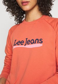 Oranžová mikina s kulatým výstřihem, zdobená námořnickým logem "Lee Jeans" a růžovým vlnitým akcentem. Měkká textura a raglánové rukávy.