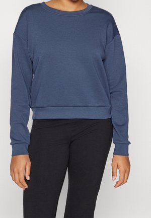 Person wearing a blue long-sleeve sweatshirt and black pants, standing against a plain light background.