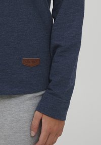 Temno modra majica z dolgimi rokavi iz teksturirane tkanine, s temno rjavim usnjenim logotipom na spodnji levi strani, kombinirana s sivo tkanino.