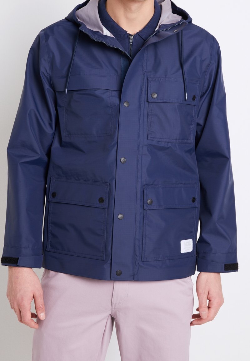 Navy blue windbreaker jacket with gray hood, snap buttons, and four outer pockets. Smooth texture, featuring a small logo patch.