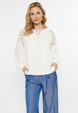 Witte blouse met kantmouwen, voorzien van een trekkoord bij de hals en gouden accenten, gecombineerd met blauwe broek met trekkoord. Zachte, lichte stof.
