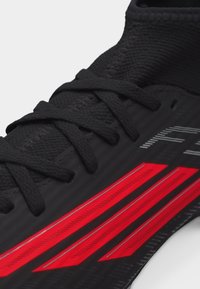 Κοντινό πλάνο μαύρου αθλητικού παπουτσιού με υφασμάτινη υφή, μαύρα κορδόνια και τρεις έντονες κόκκινες ρίγες στο πλάι κοντά στη σόλα.