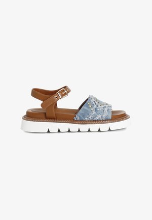 Brown leather sandal with denim upper featuring frayed accents and gold studs on a white, thick, ridged sole. Adjustable ankle strap included.