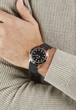 Schwarze Uhr mit rundem Zifferblatt, roségoldenen Akzenten und einem Silikonarmband, gezeigt an einer Hand, die gegen einen beigen Pullover lehnt.