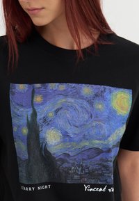 T-shirt noir avec une grande impression de "Nuit étoilée", mettant en avant des ciels bleus tourbillonnants, des étoiles jaunes et des cyprès sombres.