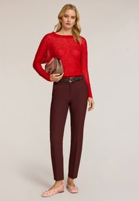 Rode doorschijnende gebreide trui, bordeaux slim-fit broek, roze flats met bloemenaccenten. Vergezeld van een bruine handtas met logodetail.