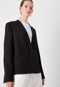 Blazer nero su misura con scollo a V, maniche lunghe e chiusura con un solo bottone. Presenta due tasche frontali con dettagli in nero.