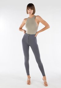 Body in tinta oliva chiaro con design aderente, abbinato a leggings grigi a vita alta con zip frontale. Sandali con tacco trasparente.