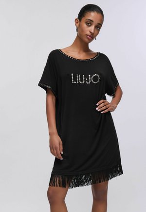 Donna con vestito nero a maniche corte con scritta "LIU JO" con borchie e orlo con frange, in piedi con una mano sul fianco su sfondo semplice.
