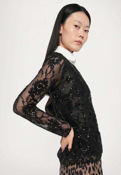 Haut noir transparent avec des broderies et des perles intriquées, manches longues et un col blanc. Accents floraux notables et tissu texturé.