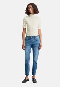 Lichtbeige kortarm turtleneck top gecombineerd met blauwe rechte jeans en zwarte instappers. De stof lijkt glad en nauwsluitend.