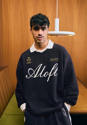 Fiatal férfi göndör hajjal és bajusszal, fekete pulóvert visel, amin az "Aloft" felirat olvasható, egy faborítású fal előtt áll, fölötte egy csillár látható.