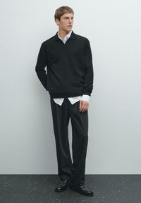 Zwarte gebreide trui met een v-hals, gedragen over een witte blouse met kraag. Gecombineerd met donkere broeken en zwarte schoenen, tegen een minimalistische achtergrond.