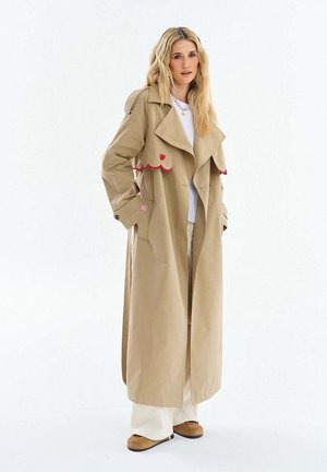 Blondynka w długim beżowym płaszczu trenczu z czerwonymi lamówkami, białej koszuli, szerokich białych spodniach i brązowych butach na jednolitym białym tle.