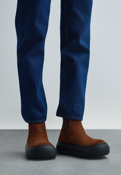 Isik, kellel on jalas pruunist nahast Chelsea saapad paksude mustade talladega ja sinised teksad, seisab hallil põrandal.