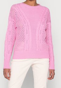 Woman wearing a light pink cable-knit sweater with open weave patterns paired with cream-colored pants against a plain background.