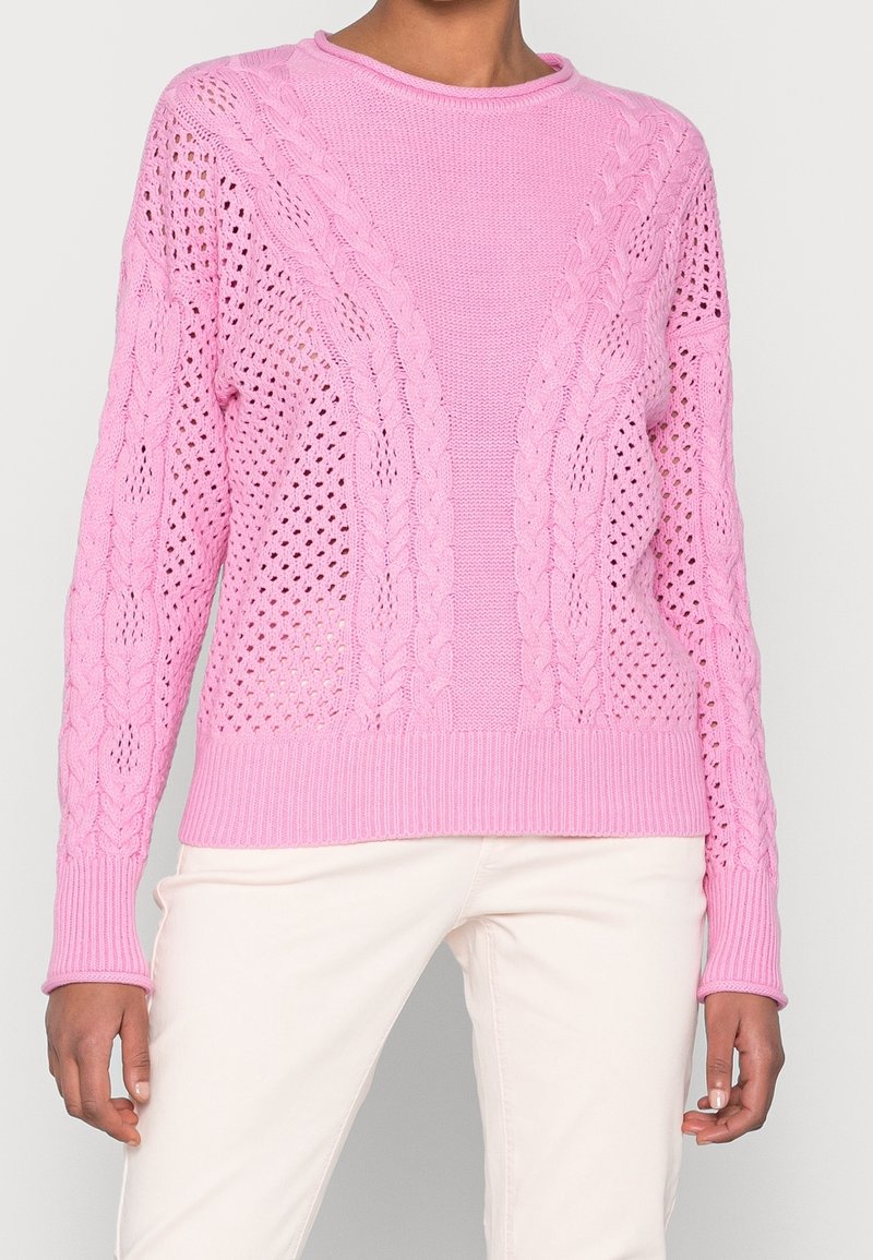Woman wearing a light pink cable-knit sweater with open weave patterns paired with cream-colored pants against a plain background.