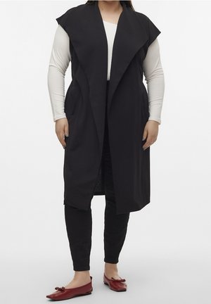 Vrouw draagt een lange zwarte mouwloze cardigan, wit shirt met lange mouwen, zwarte broek en rode ballerina's met strikjes.
