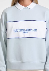 Vaaleansininen collegepaita, jossa on valkoinen vaakaraita ja sinisellä brodeerattu "GUESS JEANS 1981". Sisältää kauluskauluksen ja resorit hihansuissa.