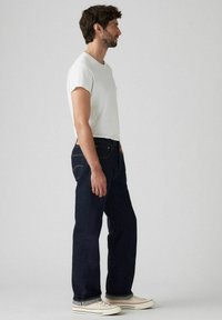 Vaqueros azul oscuro de mezclilla, con corte recto, diseño de cinco bolsillos, costuras naranjas, combinados con una camiseta blanca ajustada y zapatillas.