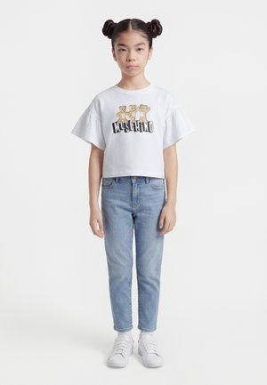 Fille avec coiffure à double chignon portant un t-shirt blanc avec des ours en peluche et le nom de la marque, un jean bleu clair, des chaussettes blanches et des baskets blanches, debout.