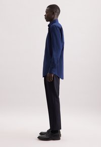 Donkerblauwe langemouwen shirt met een puntige kraag, gecombineerd met donkere broeken en zwarte schoenen. Glad materiaal met een gestructureerde pasvorm.