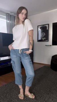 Camiseta branca cropped, calças cargo azuis claras com cintura elástica e bolsos, sandálias bege com fivela, ambiente interior com carpete cinza.