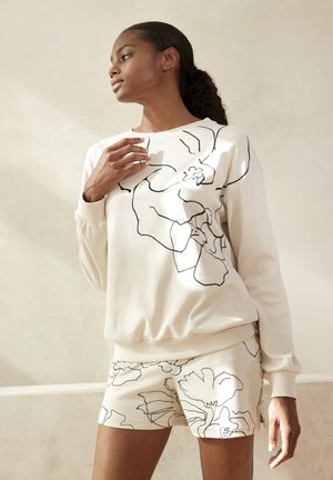 Felpa beige chiaro con design a contorno floreale nero, abbinata a pantaloni corti con lo stesso motivo. Tessuto morbido e testurizzato.